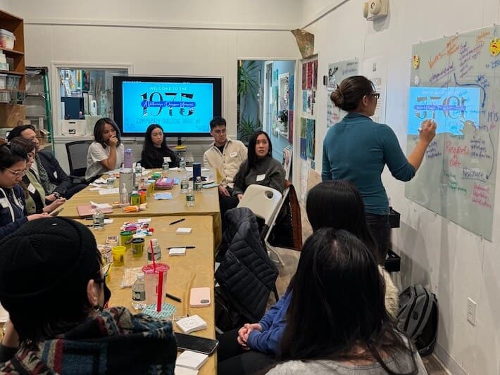 A group of people sit around a table, while one woman writes on materials posted on a wall.